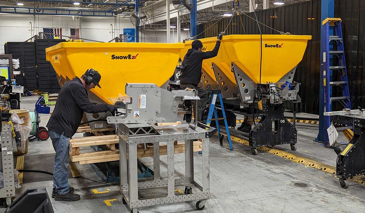 Workers in a factory assemble and inspect large yellow SnowEx snow spreaders using tools and machinery, surrounded by equipment, ladders, and pallets in an industrial environment.